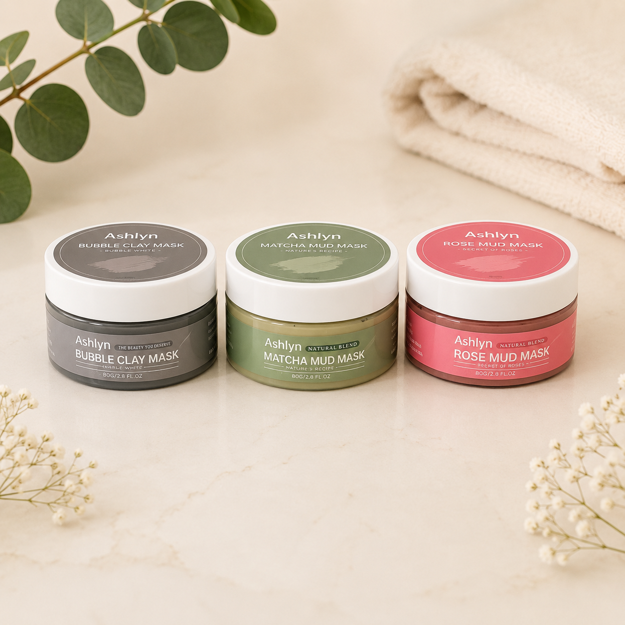 Three Ashlyn skincare mask jars arranged on a light marble surface with decorative greenery and a folded cream towel in the background. The jars are labeled Bubble Clay Mask, Matcha Mud Mask, and Rose Mud Mask.