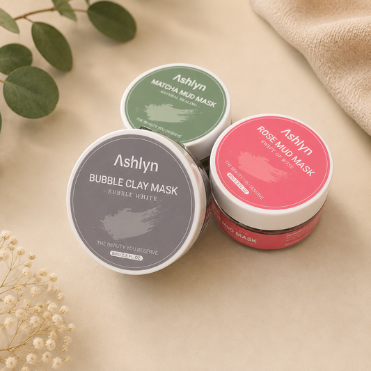 Three round cosmetic jars from Ashlyn Beauty arranged on a beige surface with decorative leaves and fabric. The jars are labeled Bubble Clay Mask (gray lid), Matdia Mud Mask (green lid), and Rose Mud Mask (pink lid).