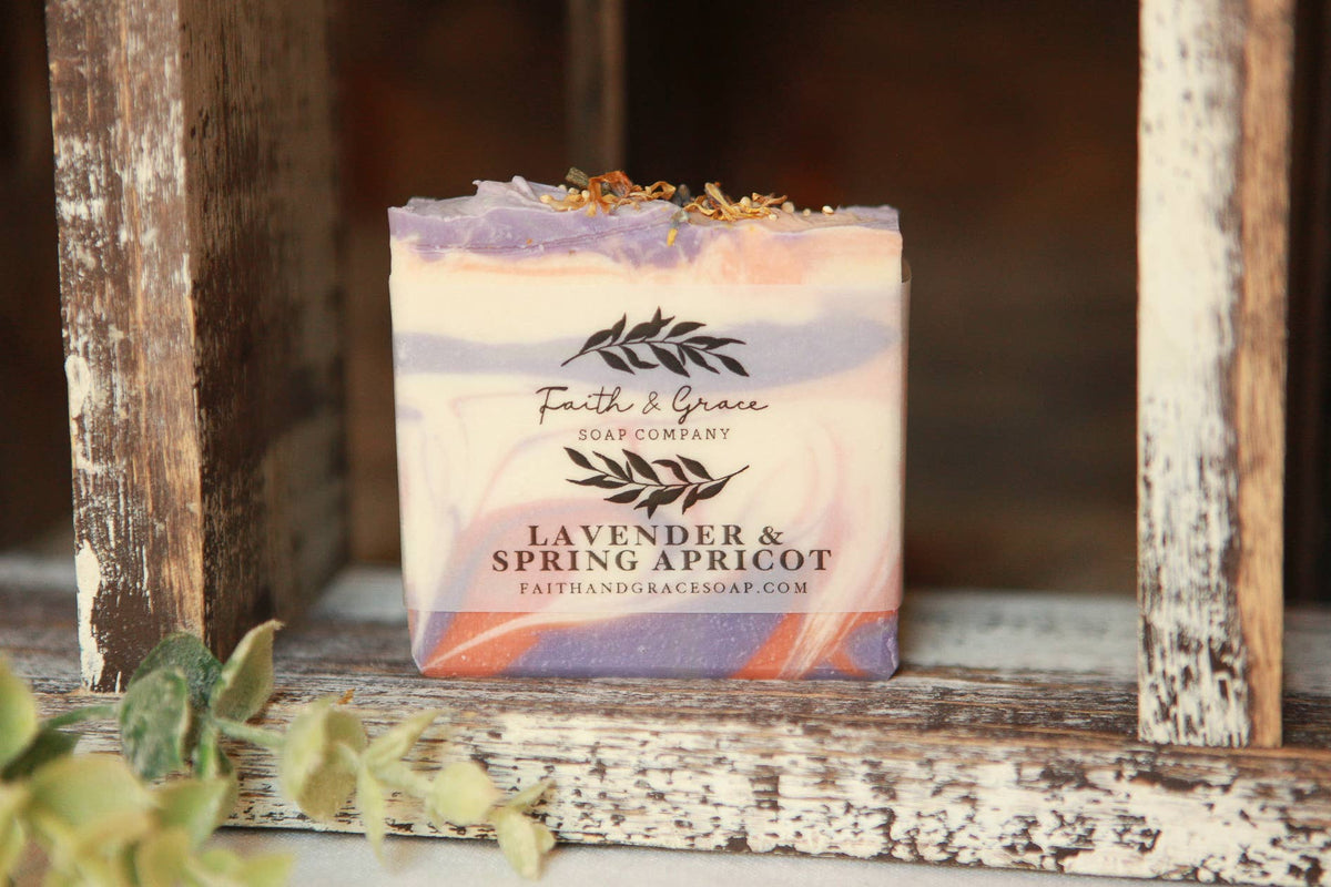 A handmade soap bar from Faith & Grace Soap Company, labeled 'Lavender & Spring Apricot,' featuring a marbled lavender, purple, peach, and orange design with dried flowers on top, displayed on a rustic wooden shelf.